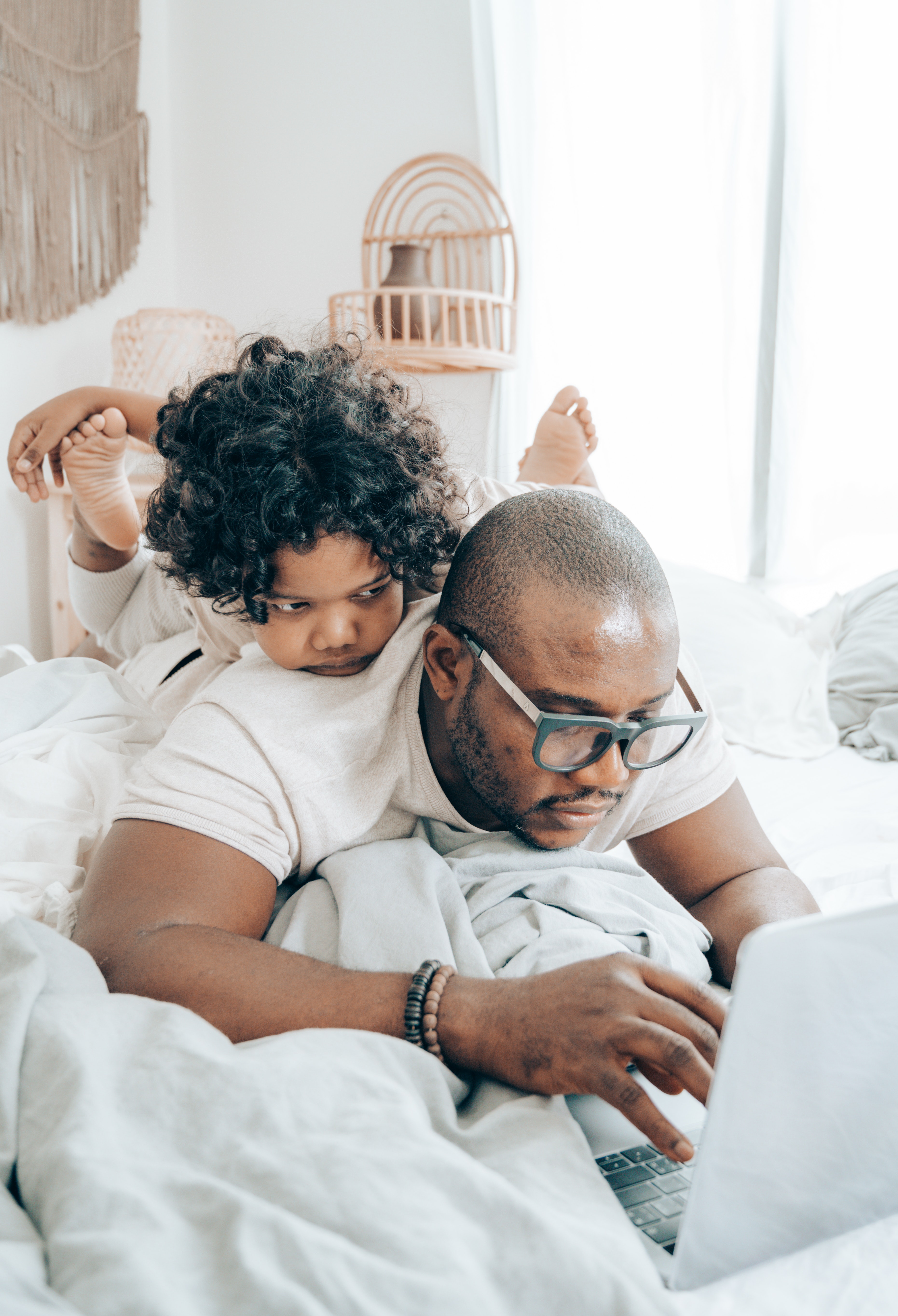 A man lays on his front working on his laptop while his child lays on his back looking over his shoulder