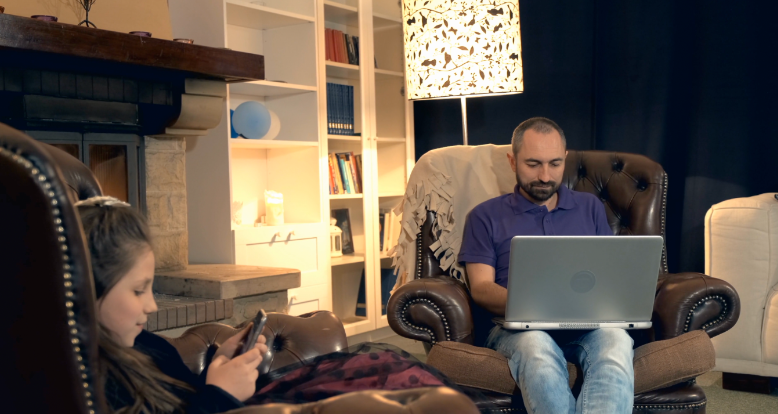 A man on a laptop and a young girl on a phone, both sitting in brown leather chairs.